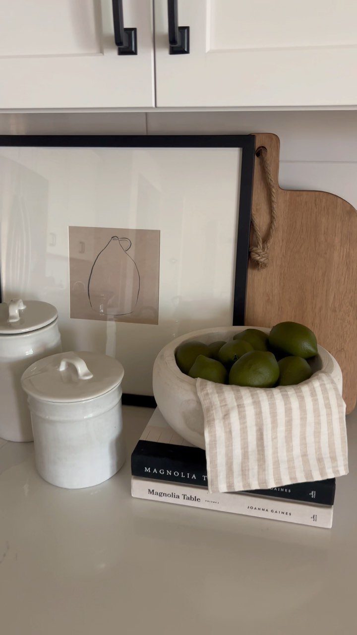 Minimalist kitchen decor featuring framed art, wooden cutting board, and limes in a white bowl.