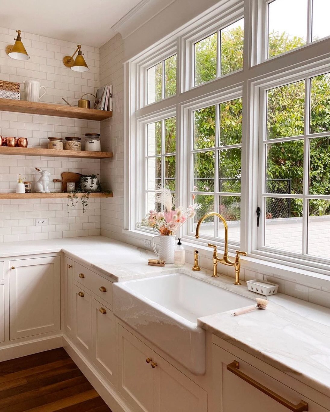 White farmhouse kitchen with an apron-front sink, marble countertops, and gold hardware by a large window.