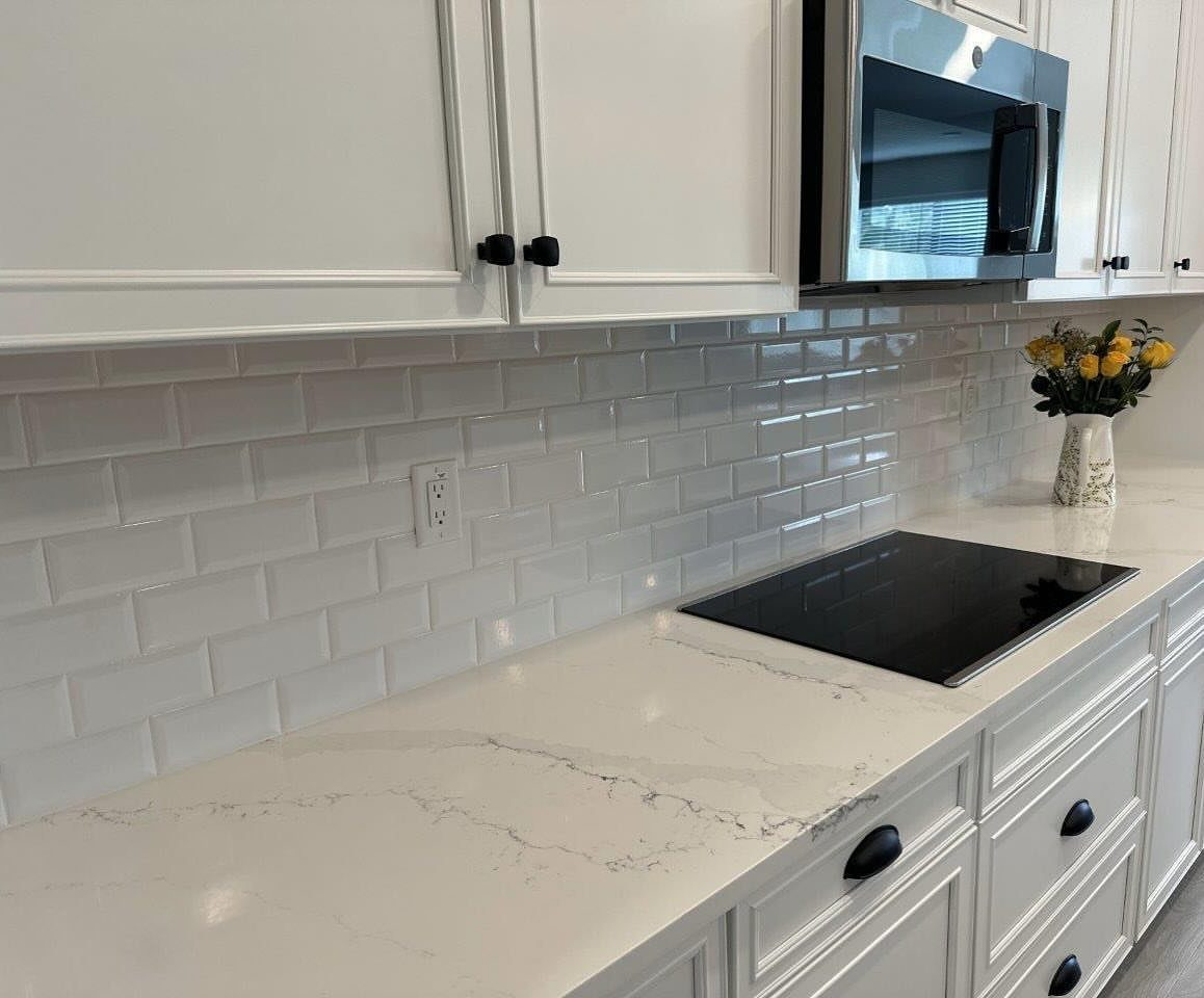 White subway tile backsplash installed in a bright kitchen with marble countertops and white cabinets.